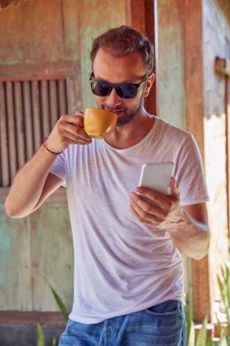 Verandada kahve/çay içerken cep telefonu kullanan adam.