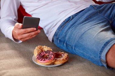 Bir teras kanepede oturan ve hücre kullanırken donut yemek adam