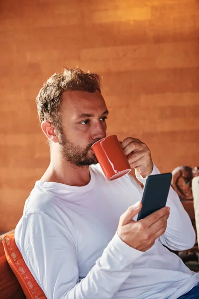Man sitting on a terrace sofa and drinking coffee/tea while usin — Stock Photo, Image