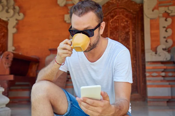 Hombre usando el teléfono celular mientras bebe café / té en el porche . — Foto de Stock