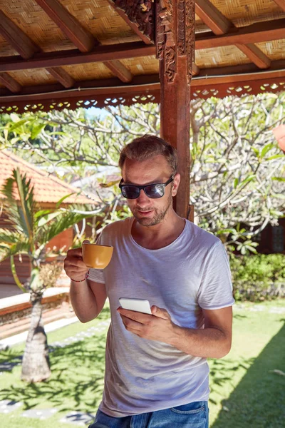 Hombre usando el teléfono celular mientras bebe café / té en el porche . — Foto de Stock