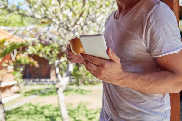 Hombre usando el teléfono celular mientras bebe café / té en el porche . —  Fotos de Stock