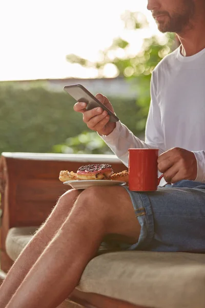 テラスのソファに座ってコーヒー/紅茶を飲みながら食べる男 — ストック写真