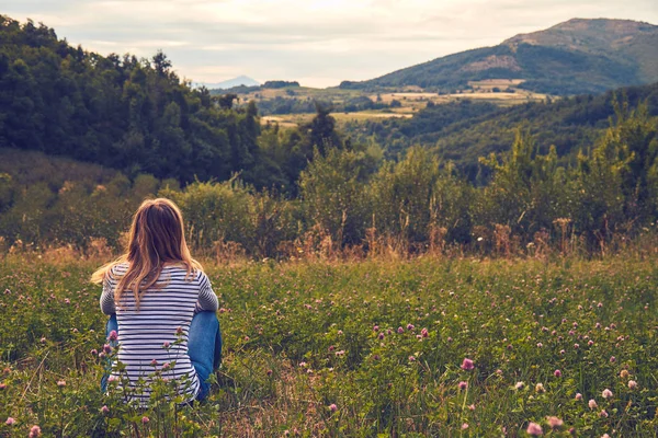 Yeşil bir çayırda oturan ve kırsal araziyi izleyen kız