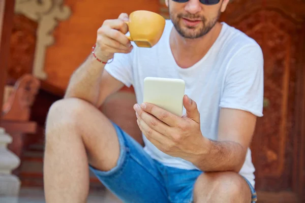 Hombre usando el teléfono celular mientras bebe café / té en el porche . — Foto de Stock