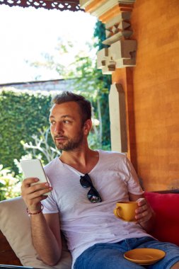 Verandada kahve/çay içerken cep telefonu kullanan adam.