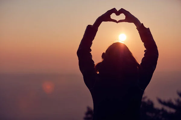 Gün batımı/gündoğumu ti elleri ile kalp şekli işareti yapan kız — Stok fotoğraf