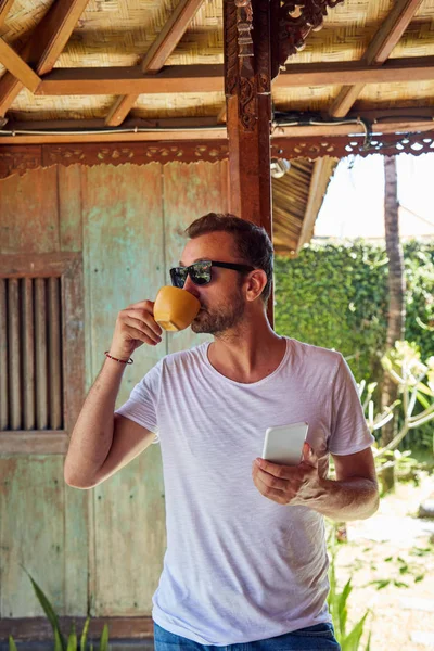 Hombre usando el teléfono celular mientras bebe café / té en el porche . — Foto de Stock