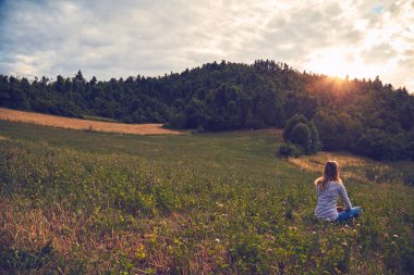 Yeşil bir çayırda oturan ve kırsal araziyi izleyen kız