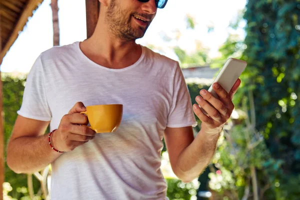 Hombre usando el teléfono celular mientras bebe café / té en el porche . — Foto de Stock