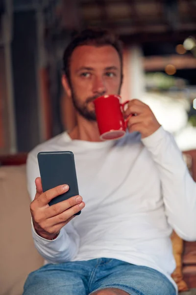 Hombre sentado en un sofá terraza y beber café / té mientras usa — Foto de Stock