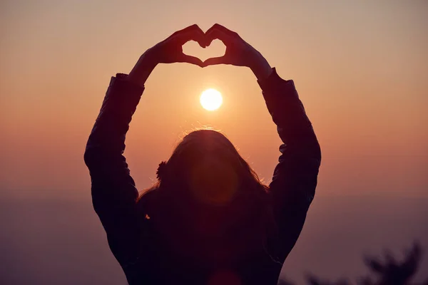 Gün batımı/gündoğumu ti elleri ile kalp şekli işareti yapan kız — Stok fotoğraf