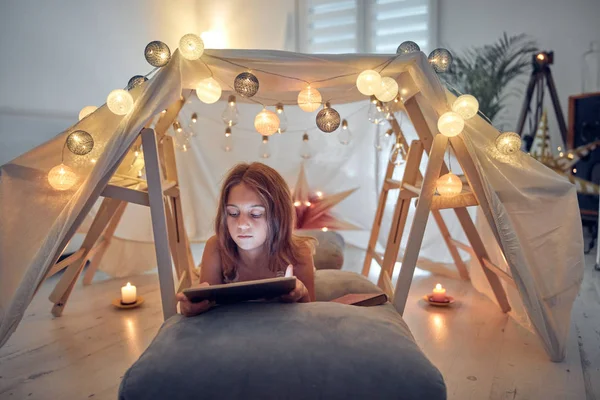 Piccola bambina di 10 anni utilizzando tablet sotto la sua tenda fatta in casa in — Foto Stock