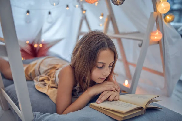 Little 10 year old girl reading classic book under her home-made — Stock Photo, Image