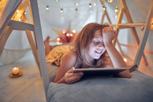 Piccola bambina di 10 anni utilizzando tablet sotto la sua tenda fatta in casa in — Foto Stock