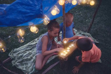 Çocuklar arka tarafında mumlar ve lampionlar olan küçük bir çadır yapıyorlar.