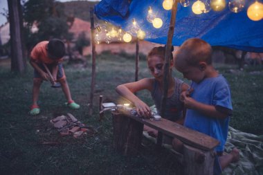 Çocuklar arka tarafında mumlar ve lampionlar olan küçük bir çadır yapıyorlar.