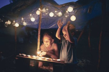 Çocuklar arka tarafında mumlar ve lampionlar olan küçük bir çadır yapıyorlar.