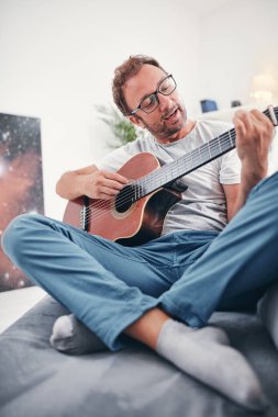 Adam oturma odasında akustik gitar çalıyor..