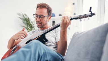 Adam oturma odasında akustik gitar çalıyor..