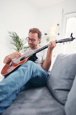 Adam oturma odasında akustik gitar çalıyor..