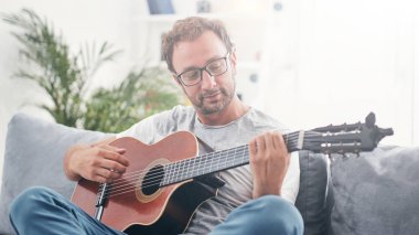 Adam oturma odasında akustik gitar çalıyor..