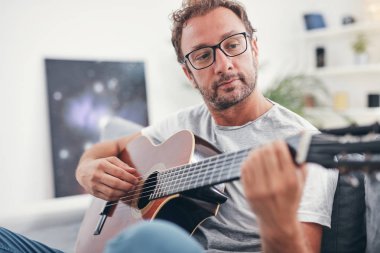 Adam oturma odasında akustik gitar çalıyor..
