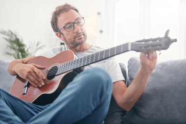 Adam oturma odasında akustik gitar çalıyor..