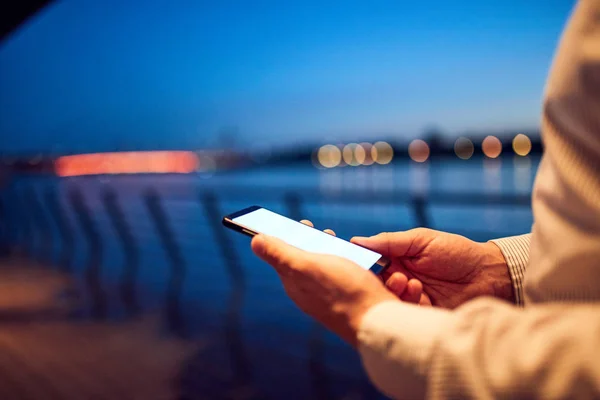 Empresário usando celular perto do rio com luzes da cidade em b — Fotografia de Stock