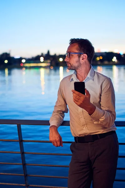 Empresario usando celular cerca del río con luces de la ciudad en b — Foto de Stock