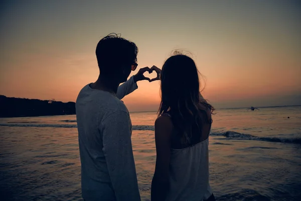 Casal fazendo símbolo de forma de coração para o amor no pôr do sol tempo sobre o oceano . — Fotografia de Stock