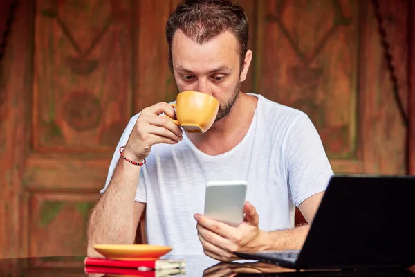 Hombre con teléfono inteligente, café / té y portátil en un porche de casa . — Foto de Stock