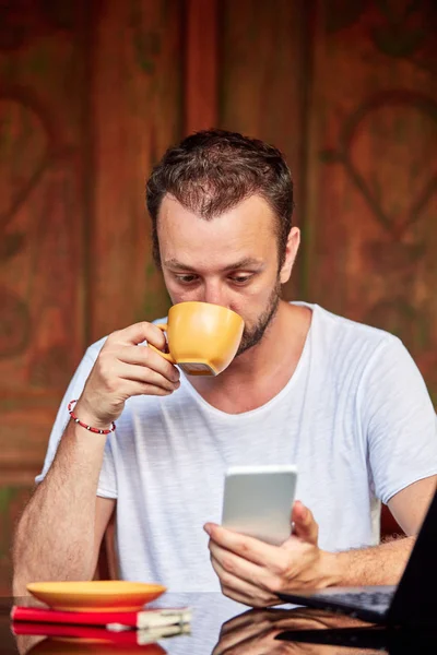 Hombre con teléfono inteligente, café / té y portátil en un porche de casa . — Foto de Stock