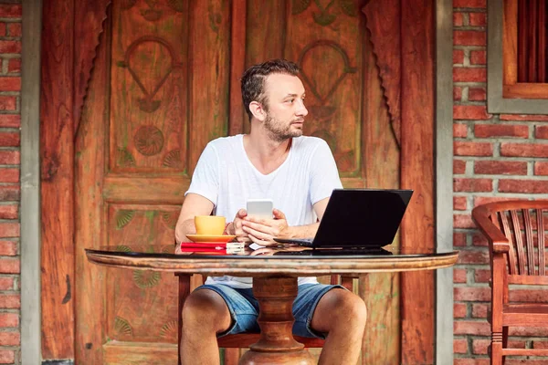 Uomo con smartphone, caffè / tè e laptop su un portico di casa . — Foto Stock
