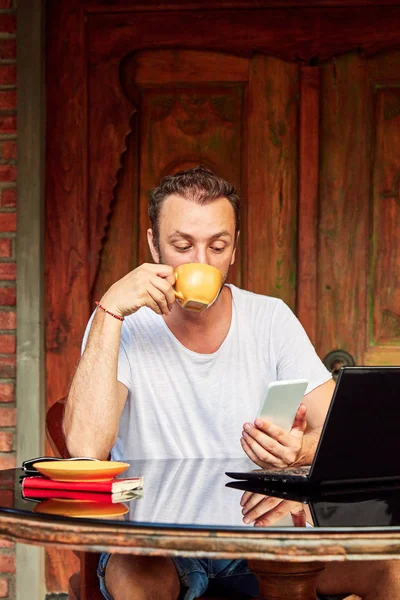 Hombre con teléfono inteligente, café / té y portátil en un porche de casa . — Foto de Stock