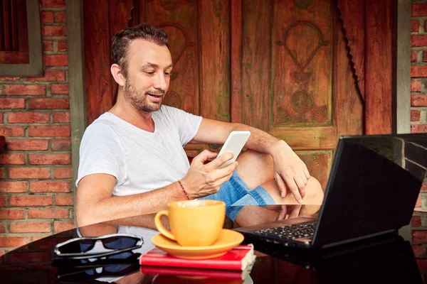 Hombre con teléfono inteligente, café / té y portátil en un porche de casa . — Foto de Stock