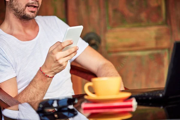 Hombre con teléfono inteligente, café / té y portátil en un porche de casa . — Foto de Stock