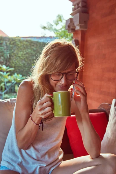 Söt kvinna med smartphone och dricka te på en veranda soffa. — Stockfoto