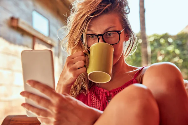 Mujer linda usando teléfono inteligente y beber té en un sofá porche . — Foto de Stock