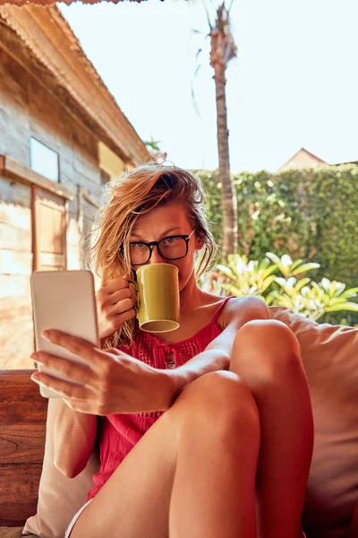 Симпатична жінка використовує смартфон і п'є чай на дивані ганку . — стокове фото