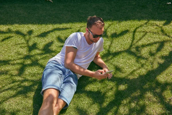 Hombre disfrutando mientras usa su teléfono inteligente en la hierba en verano —  Fotos de Stock