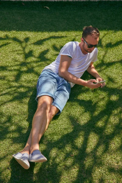 Ο άνθρωπος απολαμβάνει ενώ χρησιμοποιεί το smartphone του στο γρασίδι στο σάθερτι — Φωτογραφία Αρχείου