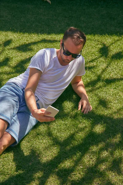 Ο άνθρωπος απολαμβάνει ενώ χρησιμοποιεί το smartphone του στο γρασίδι στο σάθερτι — Φωτογραφία Αρχείου