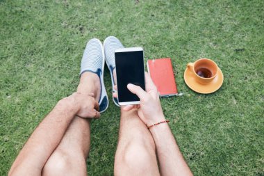 Adam akıllı telefon kullanarak ve bir çim üzerinde kahve / çay içme.