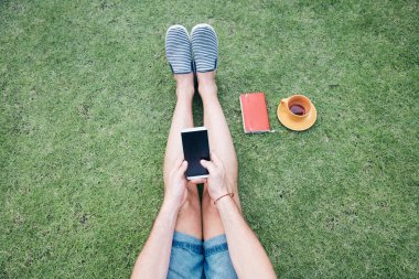 Adam akıllı telefon kullanarak ve bir çim üzerinde kahve / çay içme.