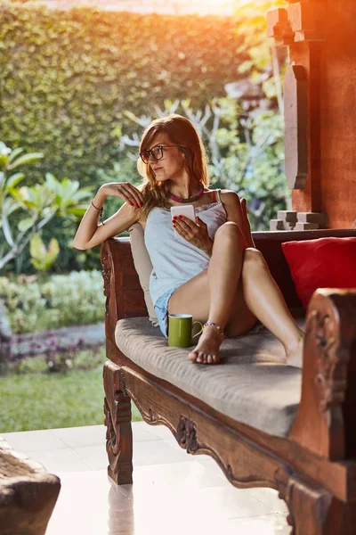 Carino donna utilizzando smartphone e bere tè su un divano portico . — Foto Stock