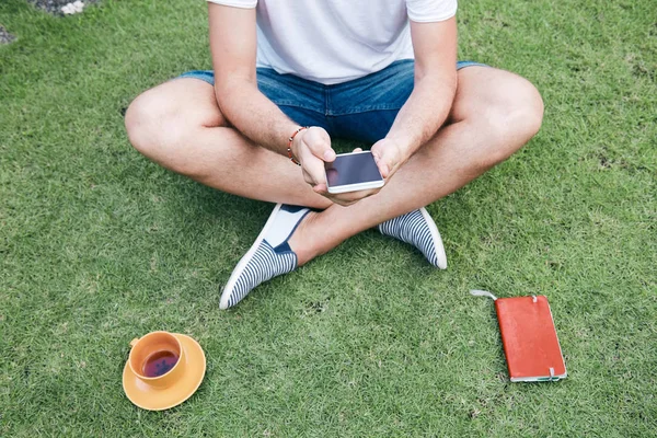 Hombre usando teléfono inteligente y beber café / té en una hierba . —  Fotos de Stock