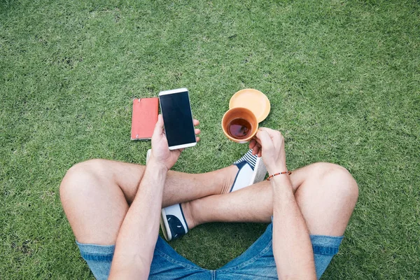 Hombre usando teléfono inteligente y beber café / té en una hierba . —  Fotos de Stock