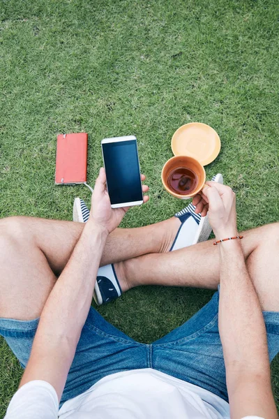 スマートフォンを使って、草の上でコーヒー/紅茶を飲む男. — ストック写真