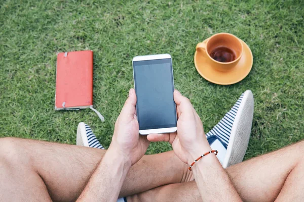 スマートフォンを使って、草の上でコーヒー/紅茶を飲む男. — ストック写真